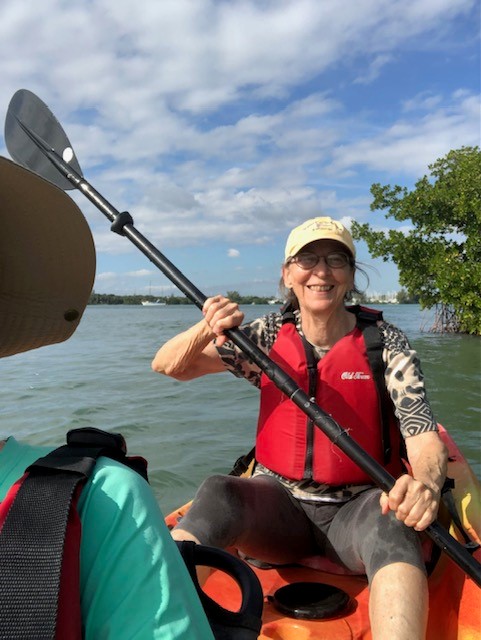 Image of Jane Birdsong paddling in a kayak. She is wearing a baseball cap, a red life jacket, and holding a paddle with one end in the water and the other out. She is smiling big.